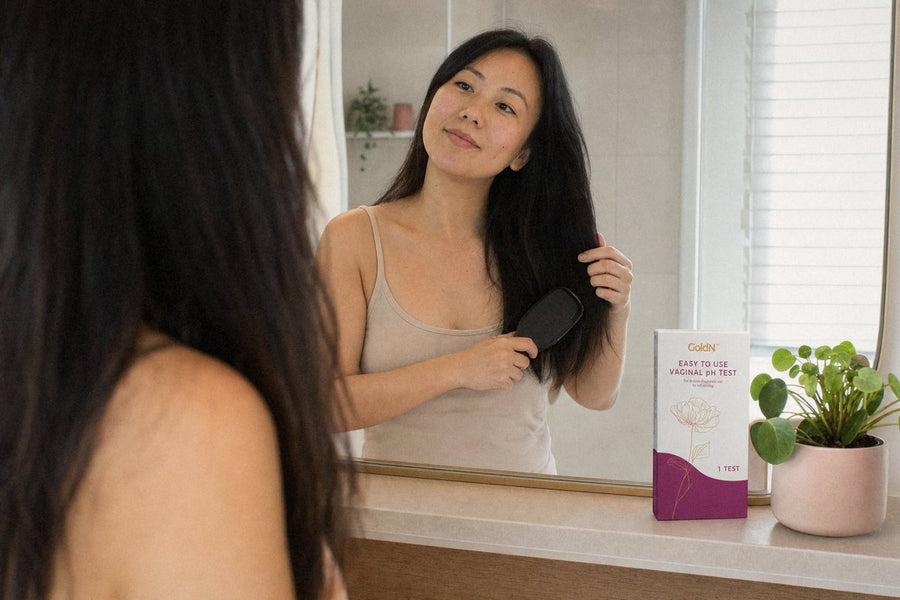 Woman brushing her hair at a bathroom mirror, GoldN Vaginal pH Test box on the counter