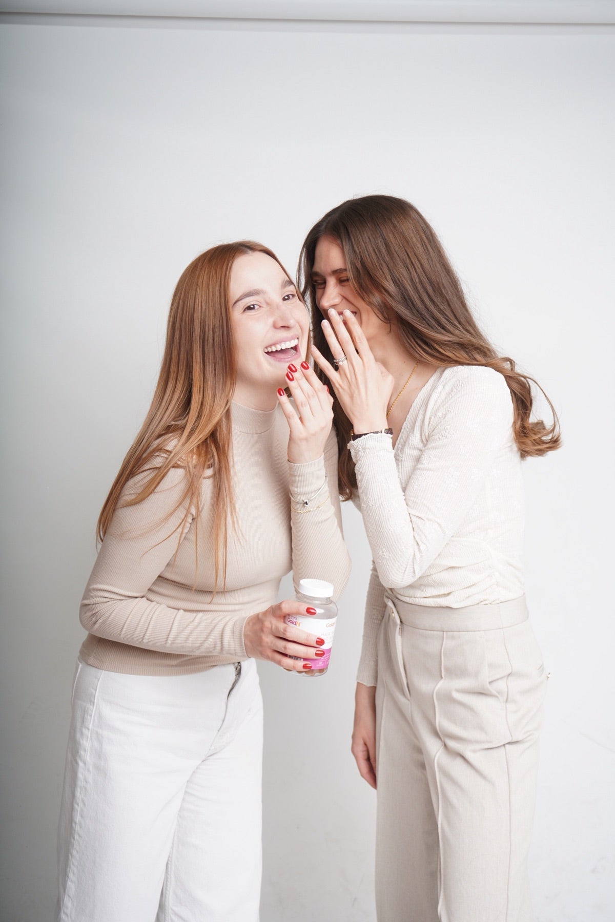 Two young women laughing in a bright studio, one holding a GoldN bottle