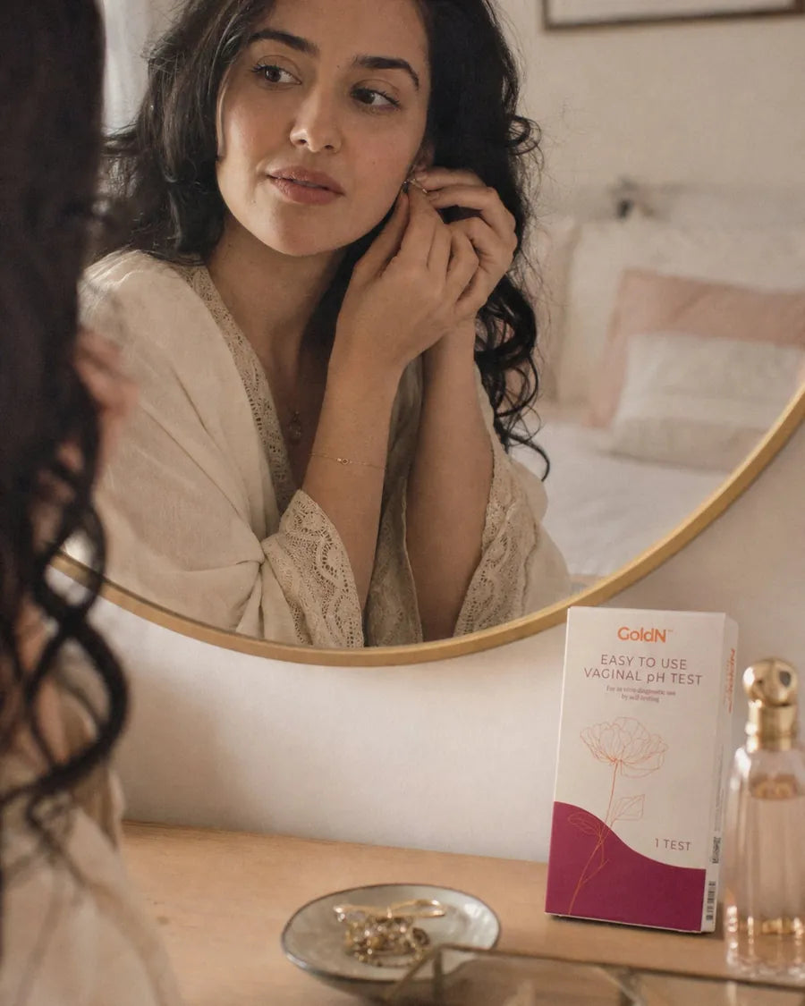 Woman putting on an earring at her dresser, a GoldN Vaginal pH Test box beside her