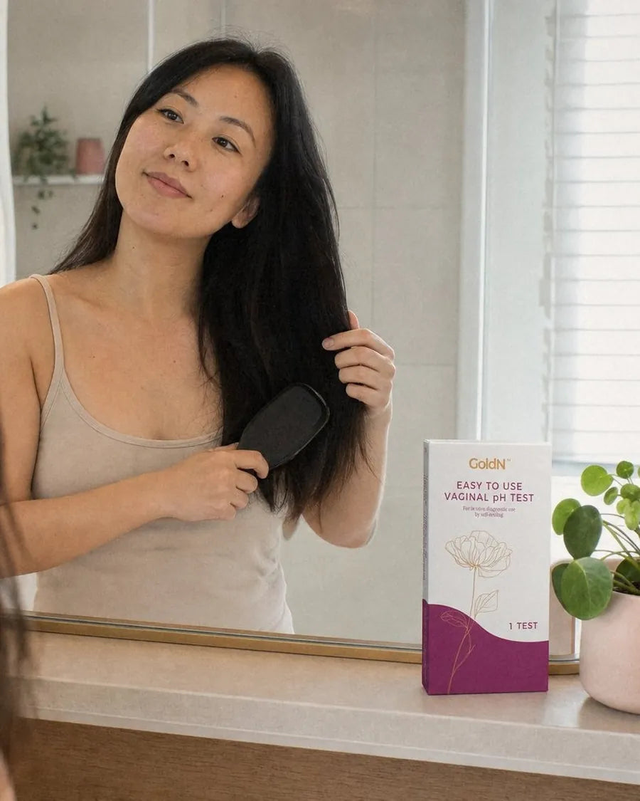 Woman brushing her hair at a bathroom vanity, a GoldN Vaginal pH Test box on the counter