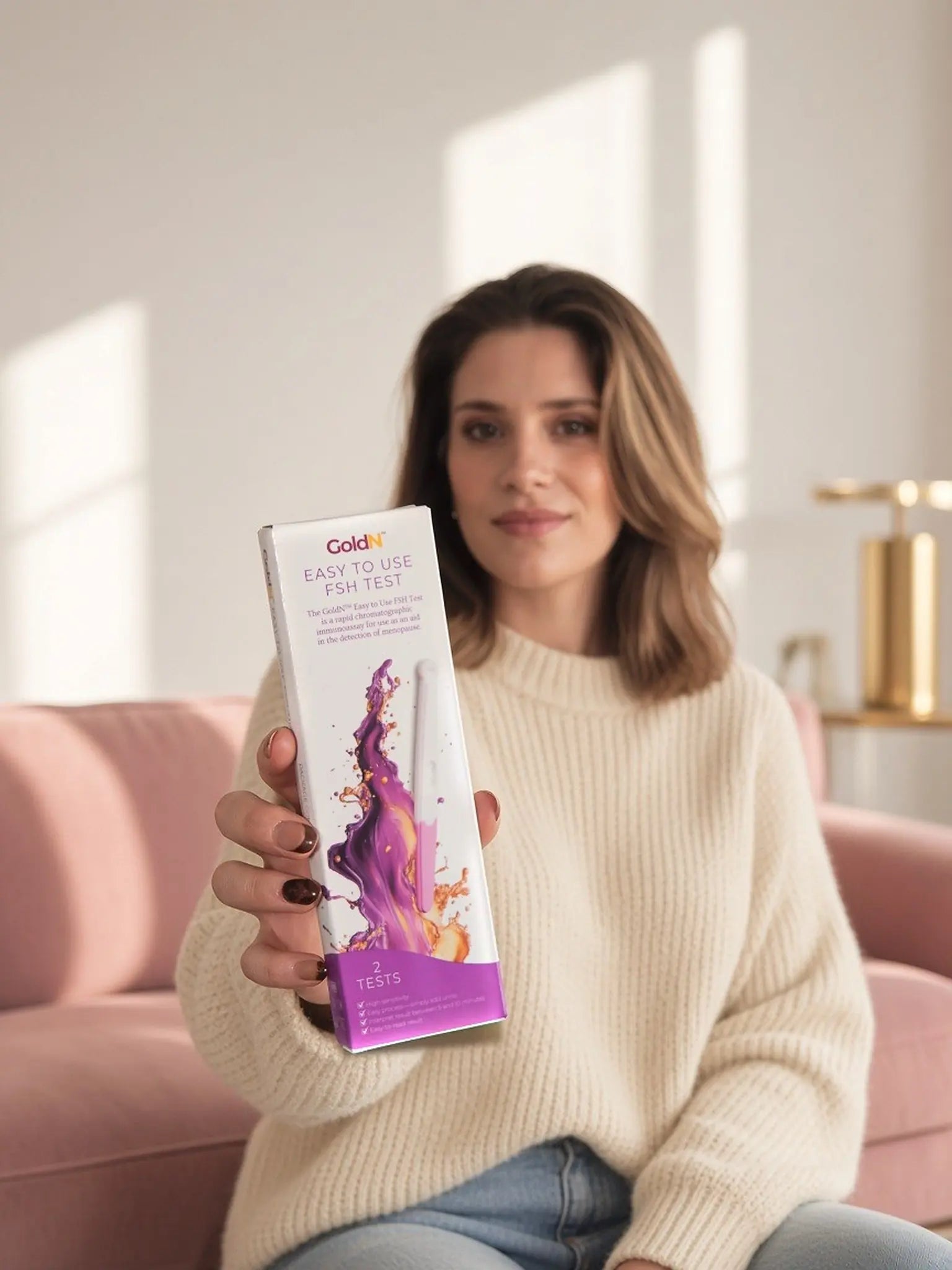 Woman on a pink sofa holding the GoldN FSH test box toward the camera