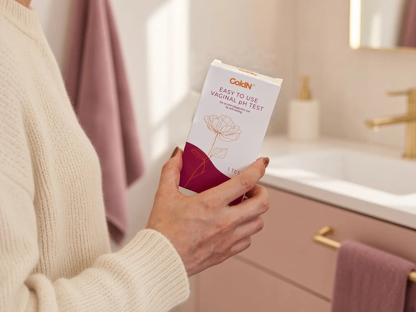 Woman in a cream sweater holding the GoldN pH test box in a pink bathroom